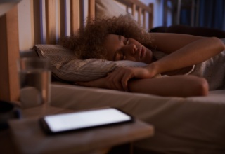 Portrait of curly-haired young woman sleeping calmly in bed at night with lit device on nightstand.
