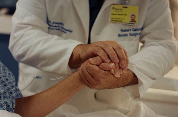 A BayCare physician wearing a white lab coat gently holds the hand of a patient lying in a hospital bed.