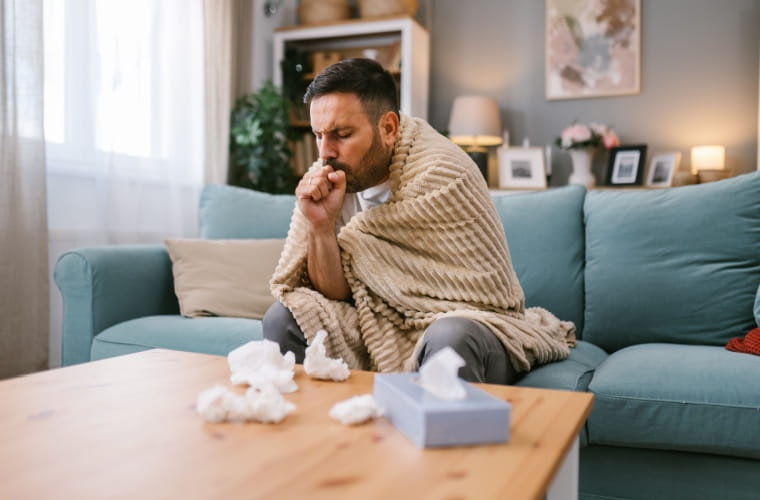 Man dealing with flu symptoms is sitting on the couch with a blanket, looking unwell, with tissues scattered around and a tissue box on the brown coffee table.