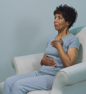 Woman sitting in chair with one hand on chest, other hand on abdomen, breathing through pursed lips.