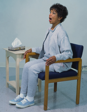 Woman sitting in chair, leaning slightly forward and coughing.
