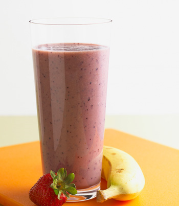 Tall glass with purple-ish colored smoothie  sitting on a table next to a banana and a strawberry.
