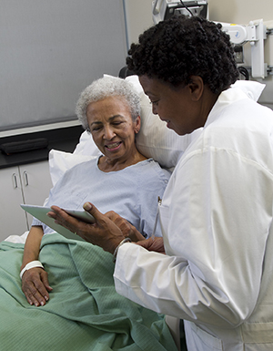 Healthcare provider with electronic tablet talking to woman in hospital bed.