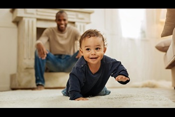 Happy darkhaired afroamerican man laughing and watching his cheerful young son crawling on the floor