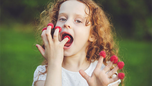 A young girl eats raspberries that she has placed on each of her fingertips.