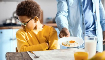 A boy turns his head away from the food his father is placing on the table.