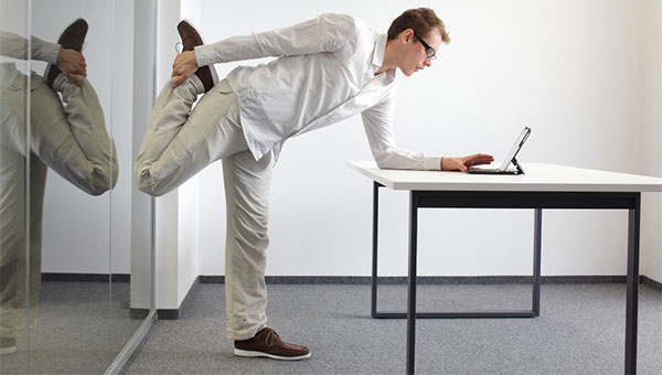 A man is standing and stretching one leg while looking at his laptop at work.