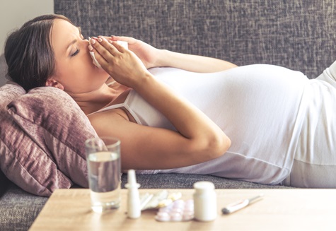 A pregnant woman is blowing her nose while resting on a sofa.
