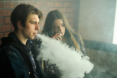 A young male exhales a vape cloud while sitting next to a young female