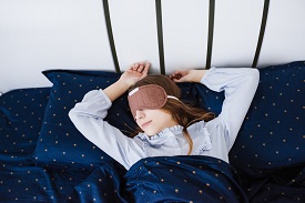 The girl sleeping and lies in her pajamas in a white linens. Blue bed. Blindfold on the head. Sleep mask