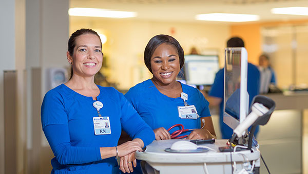 Two nurses are standing next to a computer in a hospital unit.