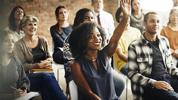 A woman sitting with a group of people and raising her hand to ask the presenter a question