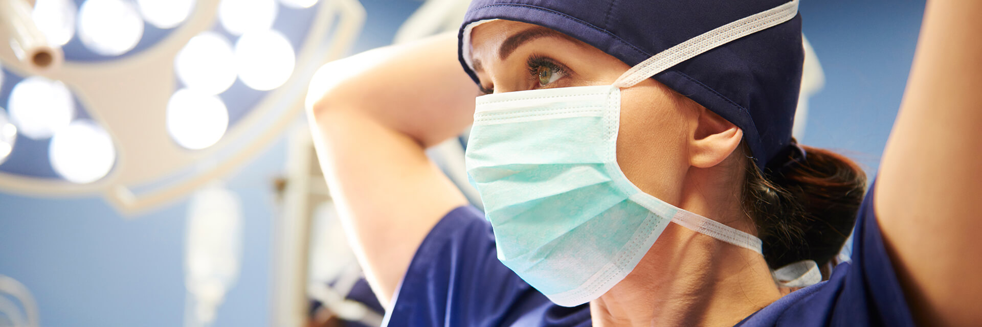 Female doctor with dark hair in blue scrub top putting on a mask