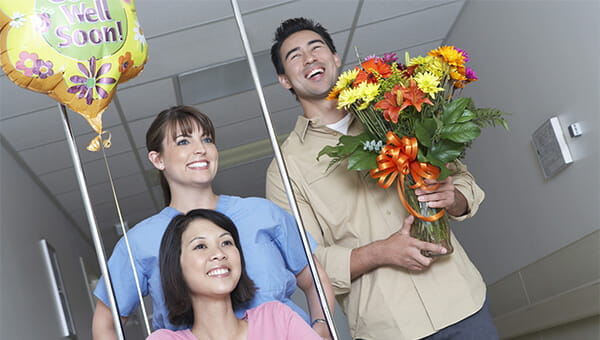 A female patient holds a balloon while a female nurse pushes her in a wheelchair and the patient's husband walks next to them holding flowers