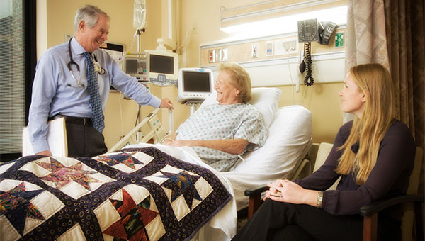 A female patient speaks with her male doctor while the patient's female visitor sits near her hospital bed