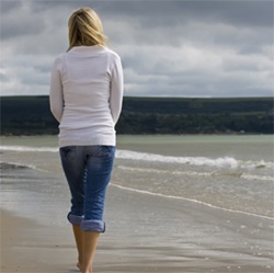 Blonde woman wearing white jacket and jeans walking down a beach shore