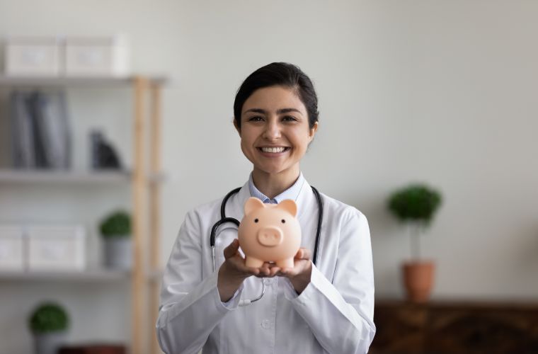 A young female physician with dark hair pulled back stands inside an office wearing a white lab coat with a stethoscope around her neck holding up a light pink piggy bank with both hands.