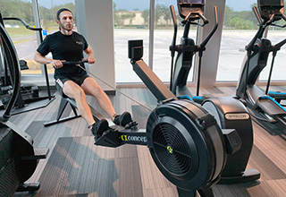 A fitness trainer, wearing a black t-shirt and black shorts, pulls back as he is working out on a rowing machine inside a fitness center, with additional fitness equipment surrounding him.