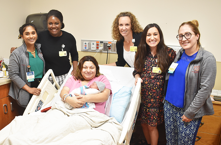 Joyful new mom with baby in her arms rests in a hospital bed, surrounded by smiling five physicians who were part of their care team. 