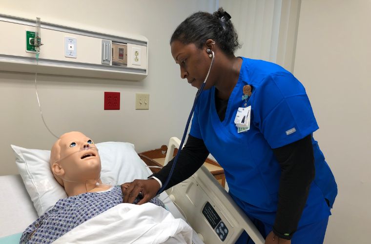 A licensed practical nurse wearing blue scrubs uses a stethoscope to hear the heartbeat of practice patient mannequin in a hospital bed during a nursing training simulation.