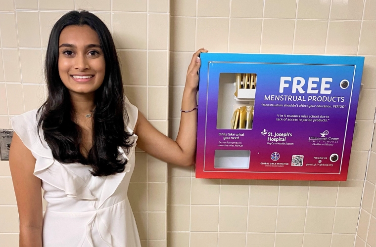 Aanya Patel, a young woman with medium length dark hair parted down the middle stands next to a feminine product dispenser in a school bathroom. She is wearing a white wrap dress with ruffles at the top. The dispenser says FREE MENSTRUAL PRODUCTS and has four logos and a QR code on it.