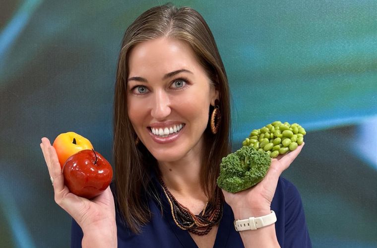 A smiling young woman with shoulder length brown holds up fruits and vegetables in both hands.
