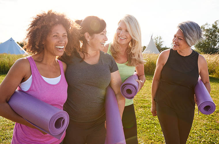 Four women outside looking at each other and smiling and holding yoga mats 