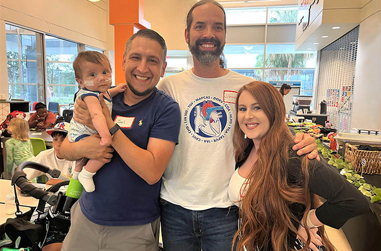 Baby who was born with heart condition is held by his father and is next to a doctor and the baby's mother. All are smiling and having fun at a heart reunion event.
