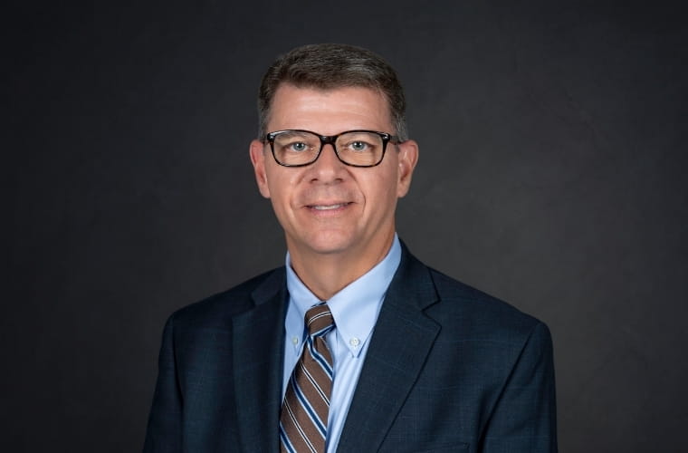 A smiling headshot of C. Todd Jones. He is wearing glasses with a dark navy suit jacket, a light blue shirt with a multi-striped tie.