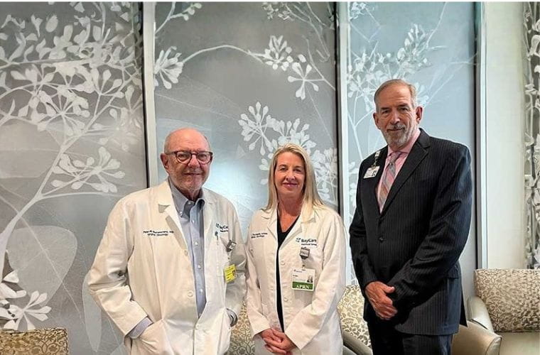 Three people -- a man and woman wearing medical coats and another man in a business suit -- stand in a lobby area in front of some windows with a design printed on them.