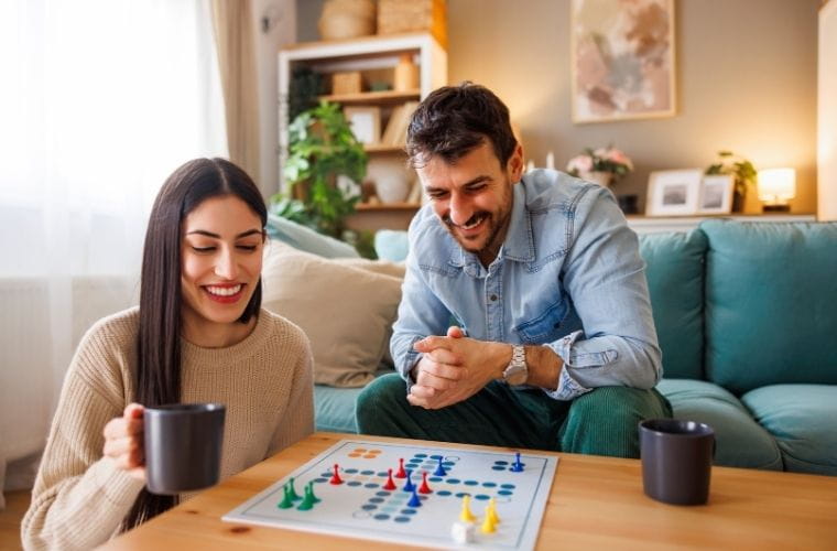 A smiling man and woman sit in a cozy living room, playing a board game with colorful pieces on a grid. A teal couch, wooden coffee table and black mugs suggest a relaxed atmosphere. A bookshelf with plants, framed art and warm lighting complete the inviting scene.