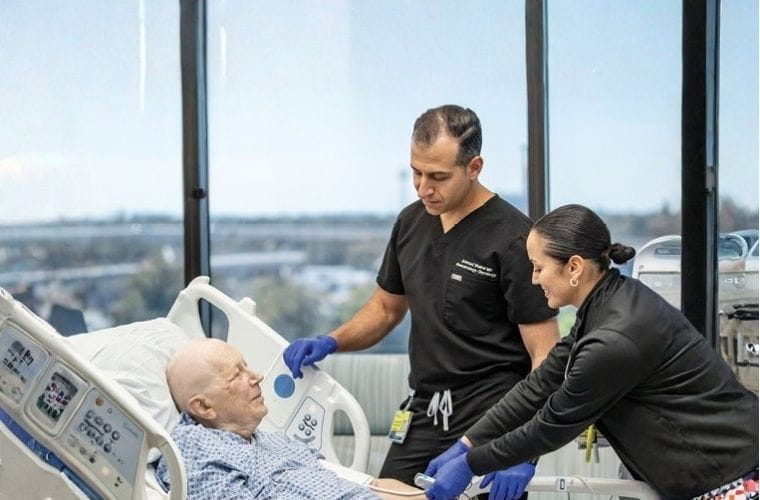 A BayCare Medical Group physician in black scrubs and a clinical nurse in blue care for an elderly patient in around a hospital bed in a hospital room with a view of St. Petersburg.