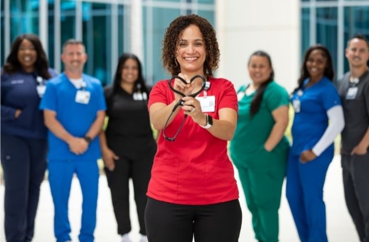 Surrounded by health care team members in scrubs of varying colors, a smiling BayCare team member holds a stethoscope in a heart shape in front of her.
