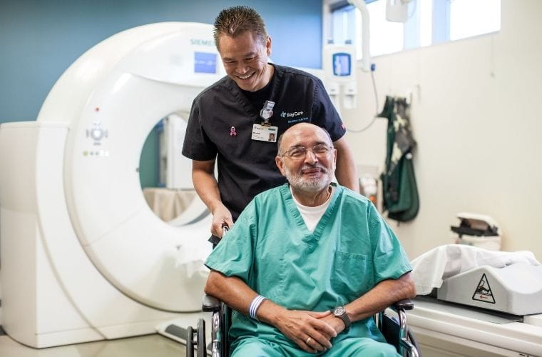 A healthcare professional wearing dark scrubs and an ID badge is standing behind a patient seated in a wheelchair. The patient is dressed in green hospital scrubs. Both are smiling. They are in a medical imaging room with a large white CT scanner in the background.