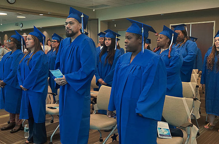 Adults dressed in blue graduation caps and gowns standing in front of their chairs at their graduation ceremony.