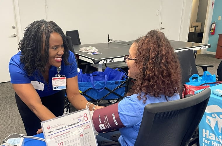 A BayCare registered nurse smiles while checking a woman’s blood pressure with a cuff during a community health outreach event. The woman sits in a chair as the nurse leans in to talk with her. A table display with information about understanding blood pressure readings sits in the foreground.