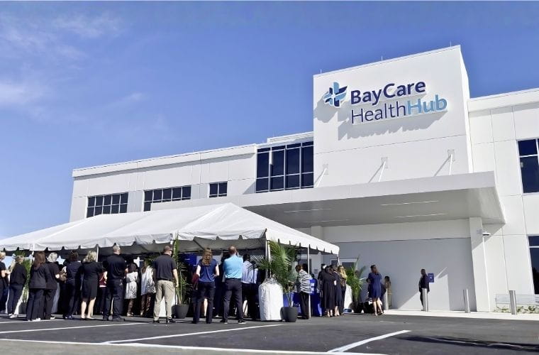 Attendees stand beneath a white event tent outside the entrance of BayCare HealthHub (Manatee) during the ribbon-cutting celebration. The front of the two-story building, featuring the BayCare HealthHub sign, is visible in the background.