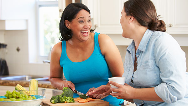 A smiling woman is preparing food while talking to her friend in the kitchen.