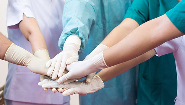 A group of doctors and nurses are standing in a circle with their right arms outstretched and their gloved right hands on top of each other