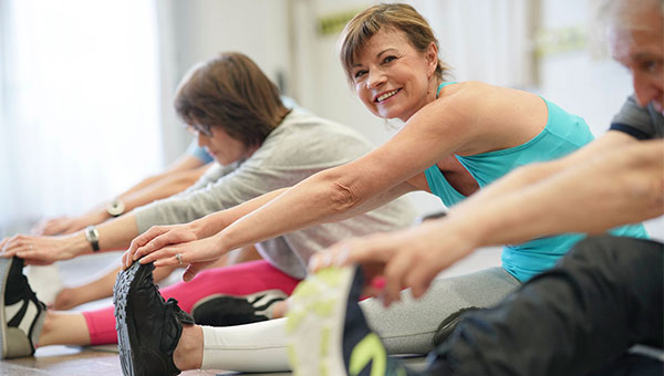 A smiling woman is sitting and stretching her legs during a fitness class.