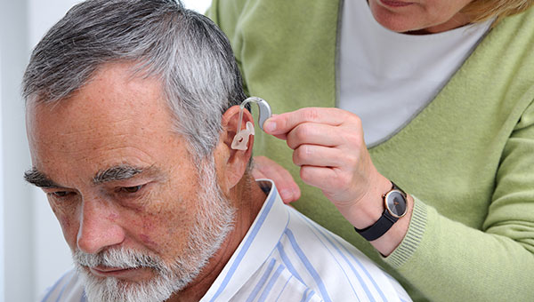 A senior man is getting a hearing aid placed in his left ear by a woman.