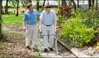 A man is walking outside next to his elderly father, who is using a walker.