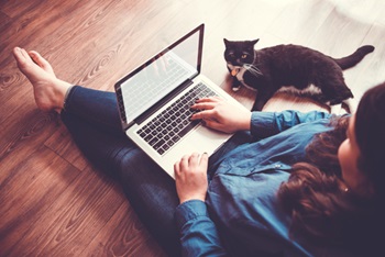 Barefoot woman sitting on floor works on laptop  while a cat watches her