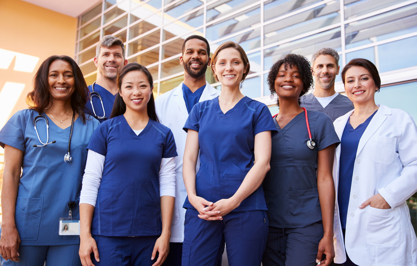 A group of diverse medical staff wearing blue scrubs standing in front of a medical building smiling.
