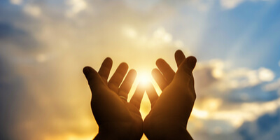 two hands praying and held up to the sky and sun