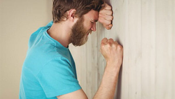 An anxious man rests his head on one hand against a wall, with the other fist clenched.