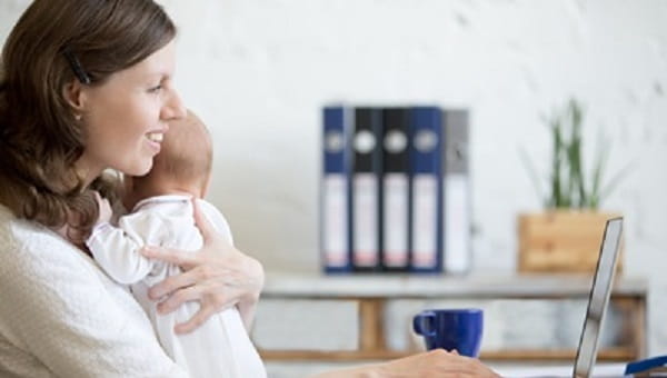 young business mom holding her newborn while working on laptop at home